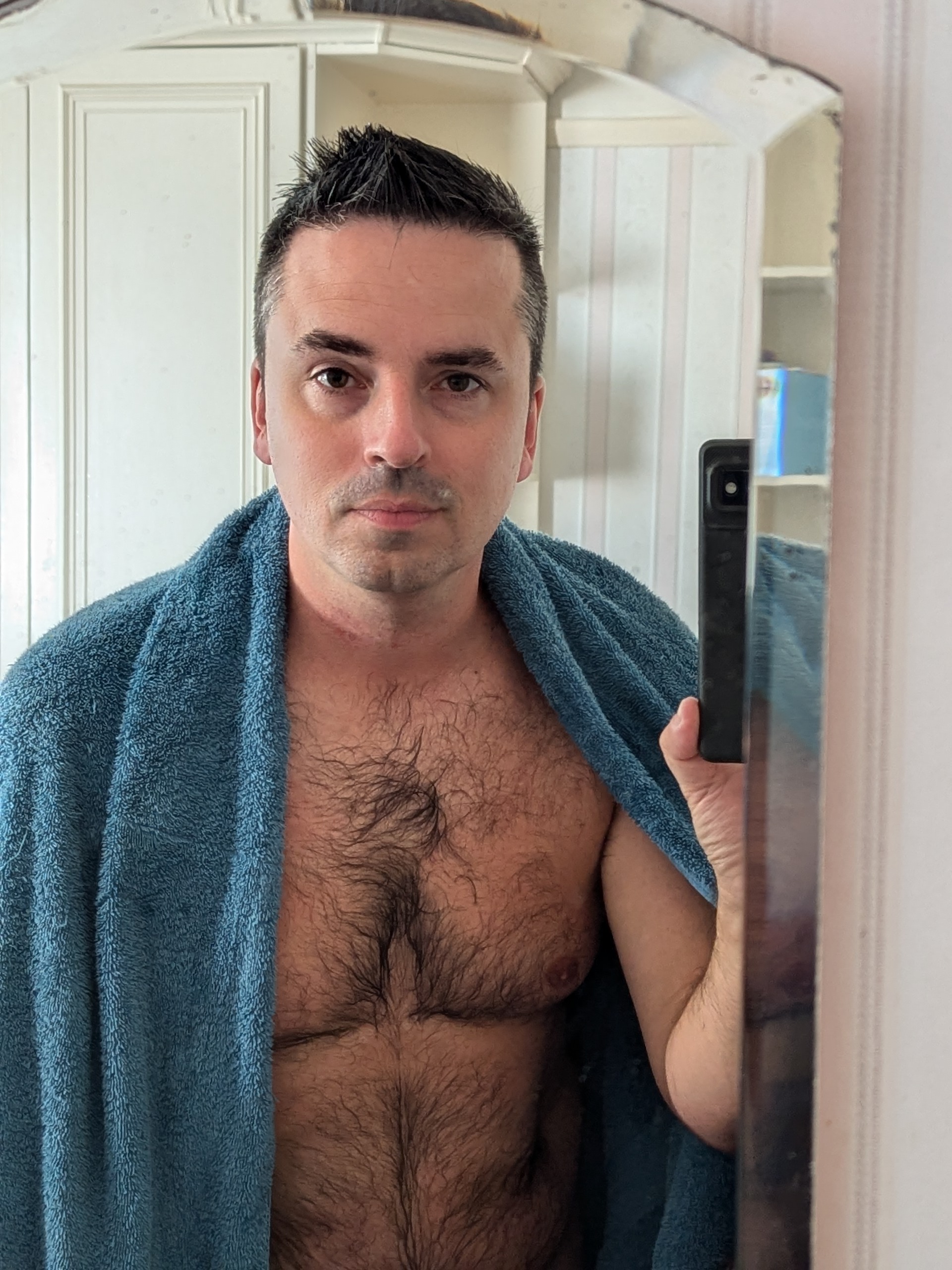 Alex standing with his chest and stomach exposed, draped with a towel and holding a razor.
