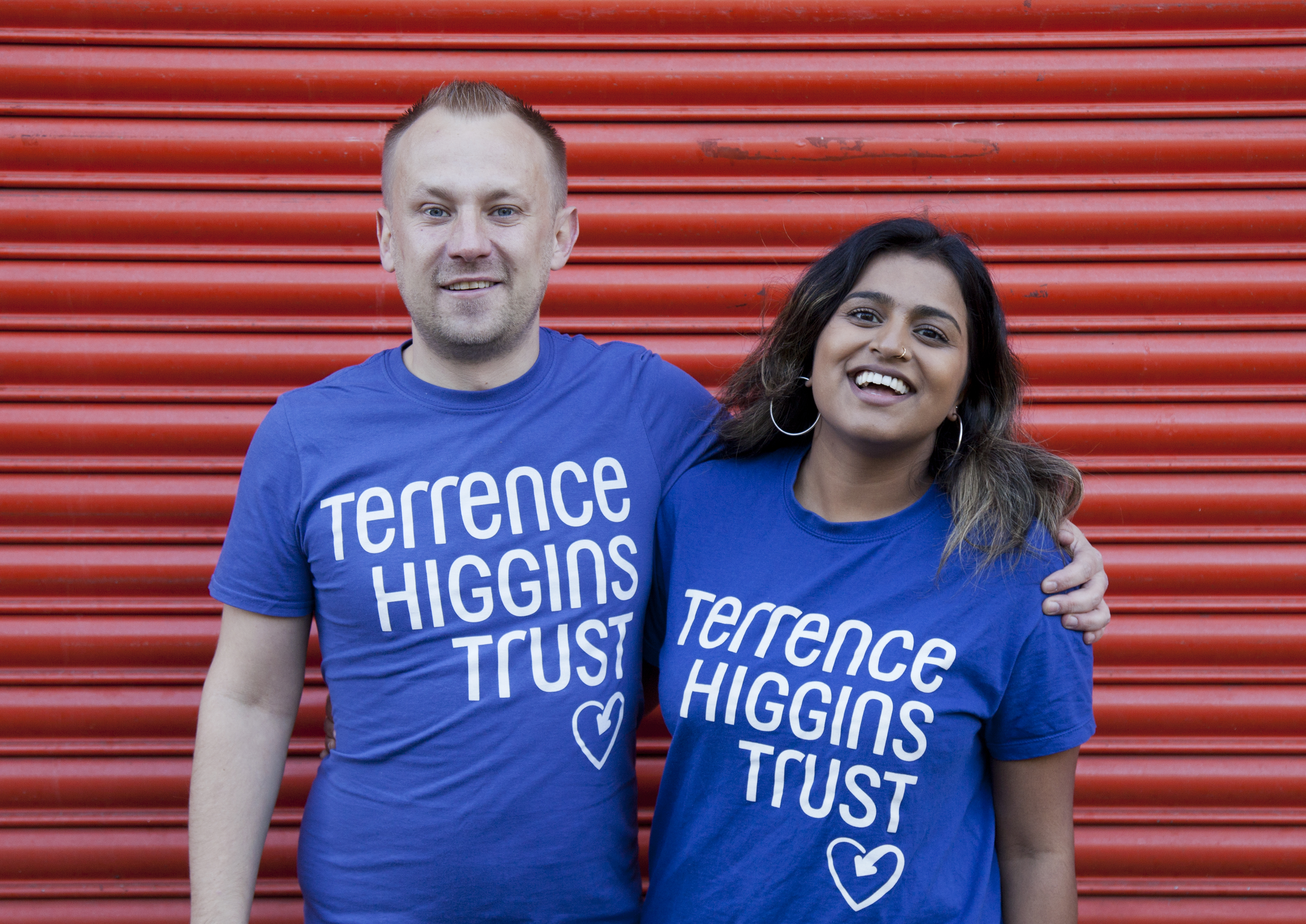 Two people wearing Terrence Higgins Trust blue T-shirts in front of red shutters 