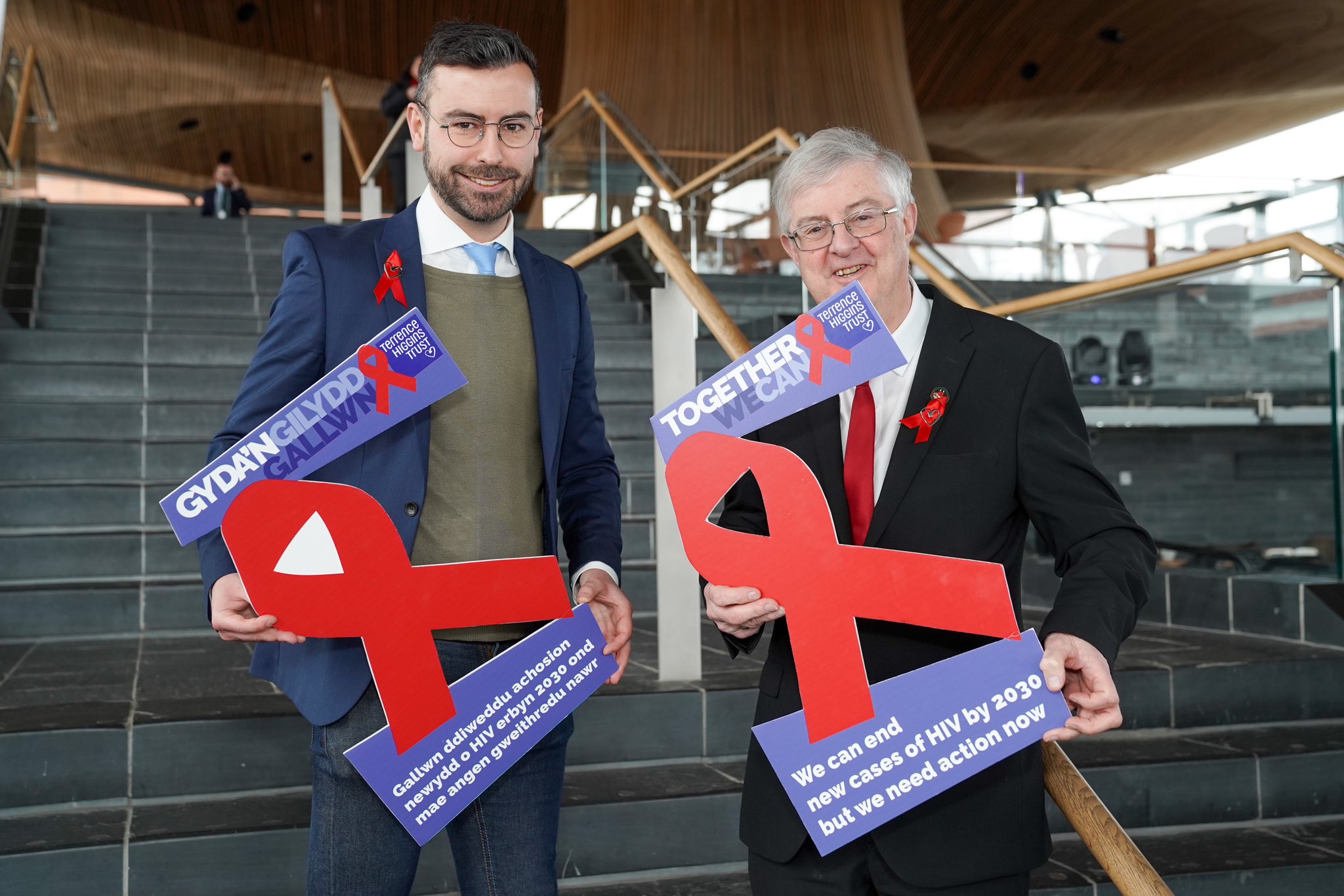 Rhys Goode and Mark Drakeford with posters saying "we can end new cases of HIV by 2030 but we need action now" in English and Welsh