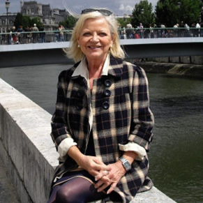 An elderly woman, smiling and sitting near a riverside.
