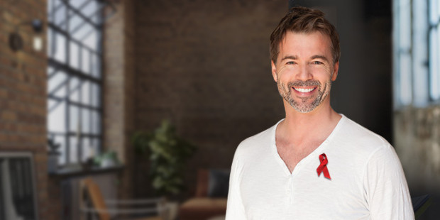 World AIDS Day A smiling man in a white shirt with a red ribbon pinned to it. The background shows a loft apartment.