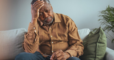 An elderly man looking down with a hand on his head.