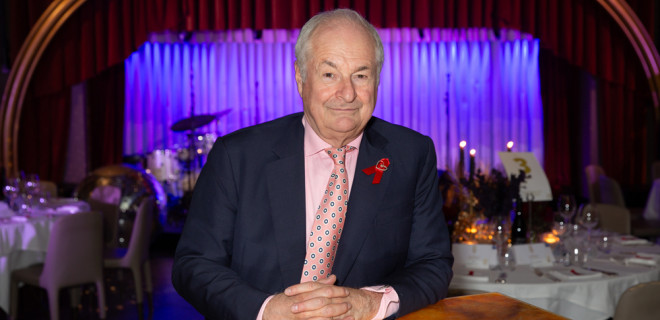 Paul Gambacini with red ribbon sat at desk at An Evening Of Radio Magic venue